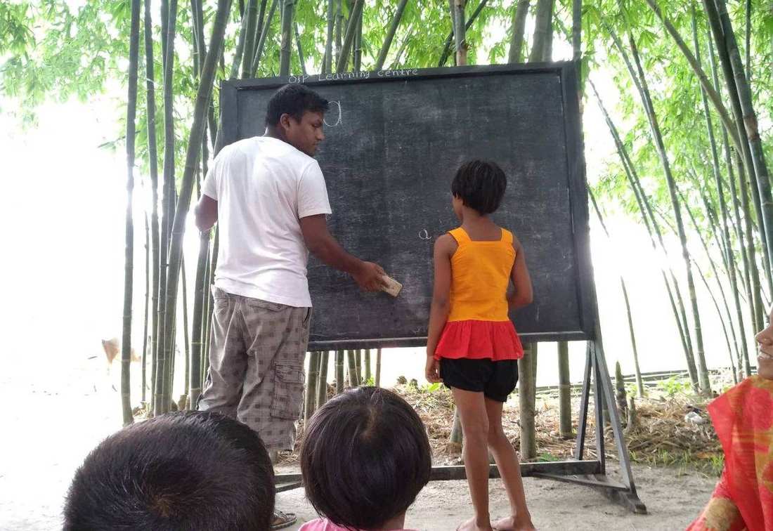 A teacher and a girl at the OfP Learning Centre blackboard under bamboo, with children seated